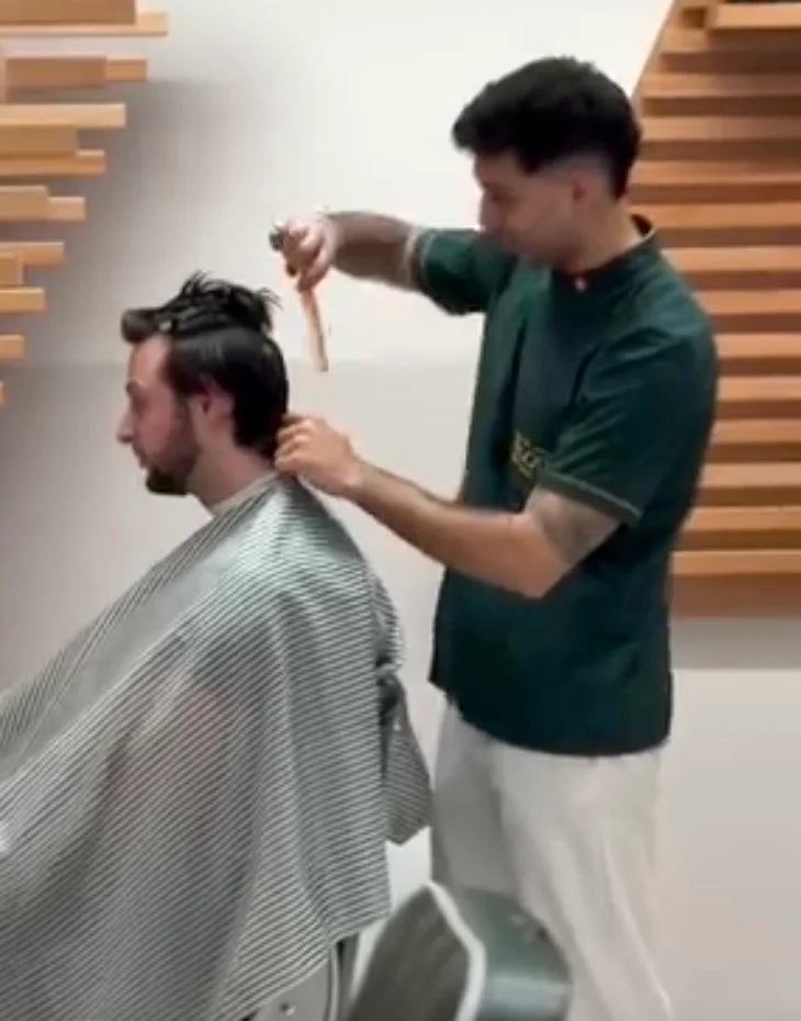 A barber working with scissors and a comb on a seated client’s medium-length hair.