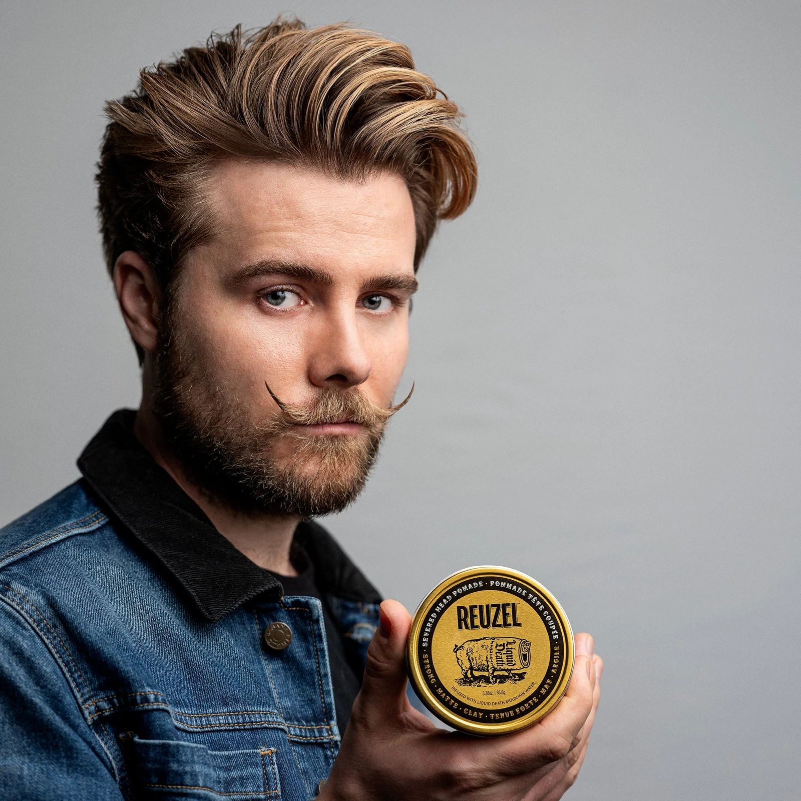 Reuzel - Strong Matte Clay - Liquid Death: Man with styled hair and a mustache holding a puck of REUZEL x Liquid Death Severed Head Strong Matte Clay Pomad