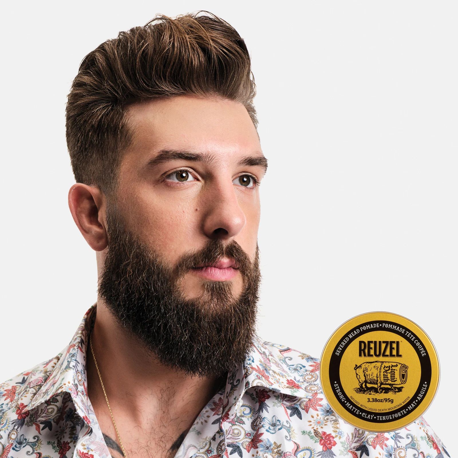 Reuzel - Strong Matte Clay - Liquid Death: Man with styled hair and a beard next to a puck of REUZEL x Liquid Death Severed Head Strong Matte Clay Pomad on