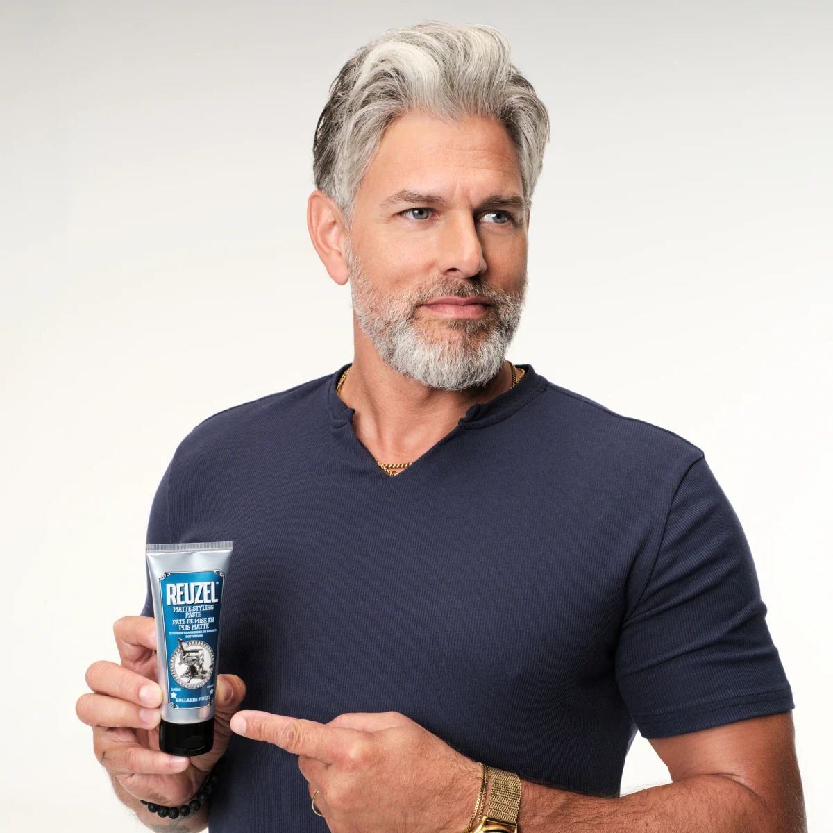 Man with grey swept back textured hair holding a tube of Reuzel Matte Styling Paste